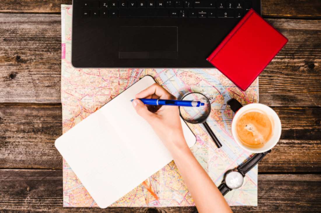 Student writing notes in a notebook with a map, laptop, coffee, and passport on a desk, representing study planning and SOP writing process.